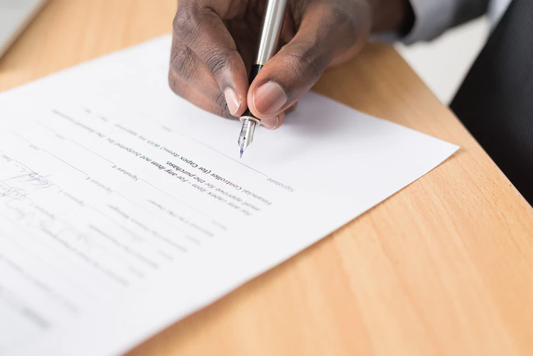 A imagem mostra um contrato sendo assinado, e uma mão segurando uma caneta de ponta fina próxima à linha de assinatura do documento.