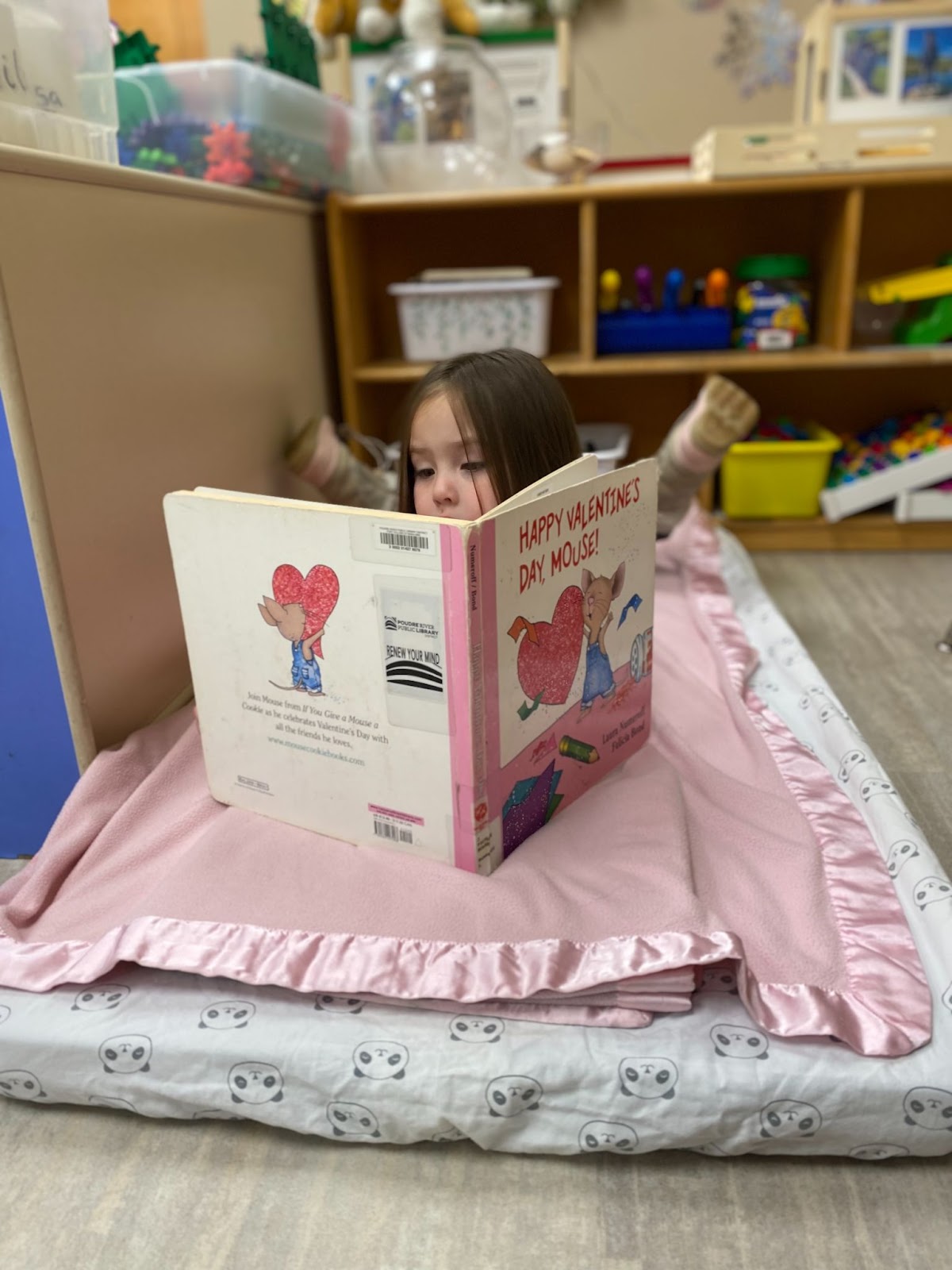 A little girl lays on a pink blanket while reading the book “Happy Valentine’s Day Mouse!”
