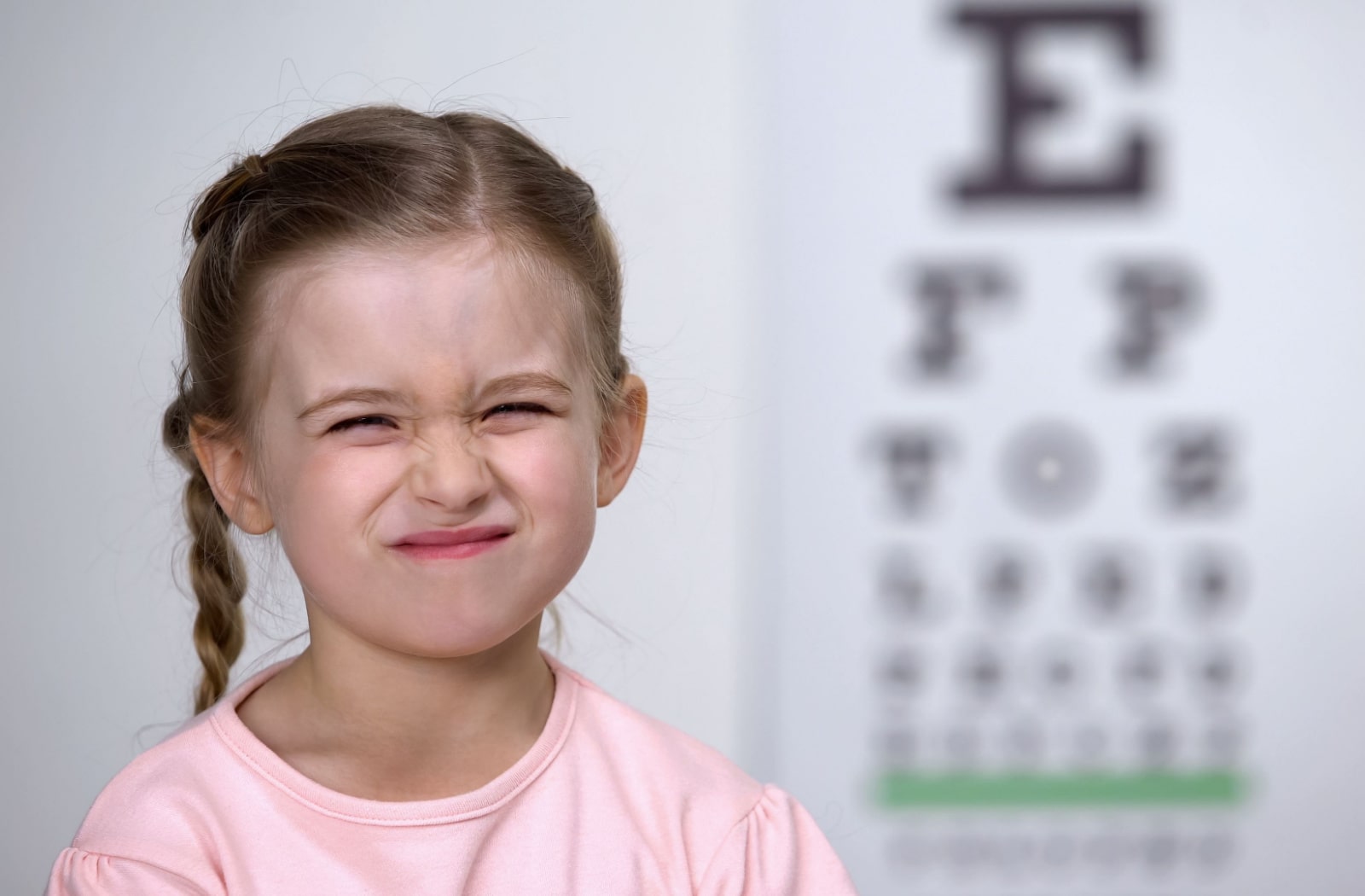A young girl standing in front of a Snellen eye chart that's out of focus, and the girl is squinting. Squinting may be a sign that your child has developed myopia.