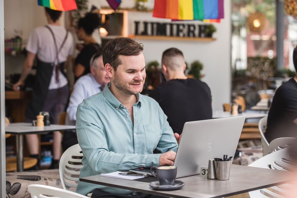 James Hill sits at a table with a laptop. In the background, the Lumiere Cafe sign and a rainbow flag are visible, as well as a few customers at tables.
