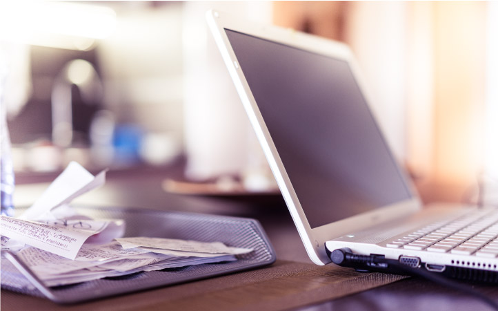 A laptop sitting open on a desk beside a metal basket filled with receipts. The image conveys the concept of digitizing your papers.