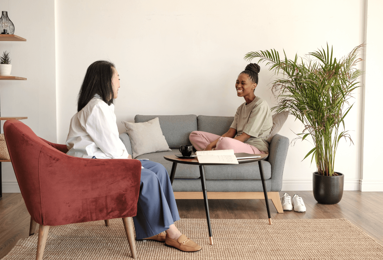 A young woman in cognitive behavioral therapy is sitting on a couch cross-legged in her therapist's office. She is smiling and looking at her counselor who is sitting in a chair near her. The image is light and looks as if the the women are happy. 