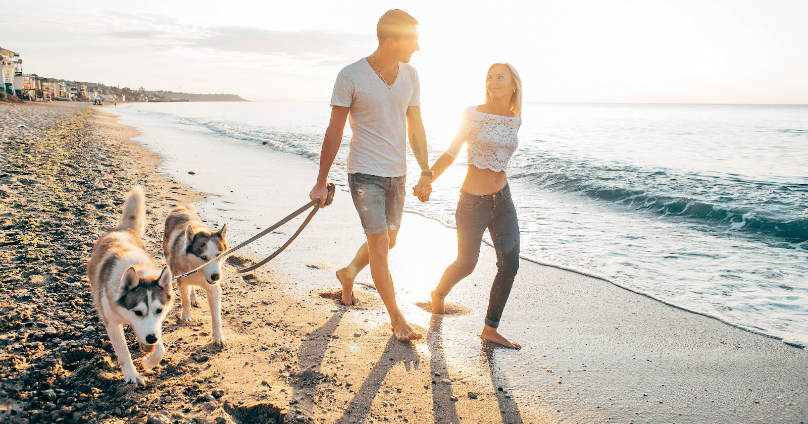 Couple walking hand in hand along beach with two huskies