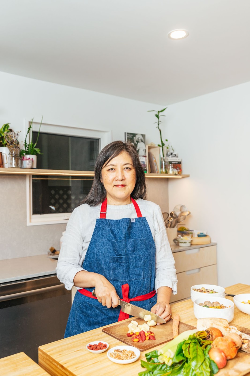 Reclaiming Chinese postpartum traditions with Feeding Mama. Image of Mona Stilwell, founder of Feeding Mama in a kitchen wearing an apron, and holding a knife to a cutting board with ingredients found in Feeding Mama dishes.