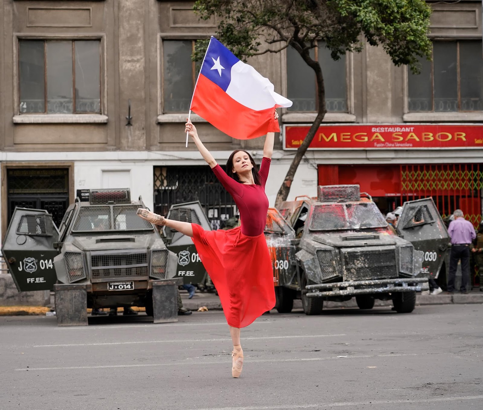 A foto mostra uma bailarina clássica segurando uma bandeira do Chile,  dançando em frente a veículos da polícia militar. 