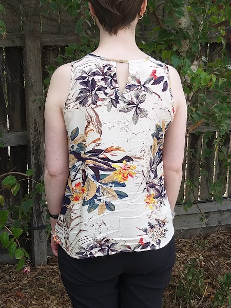 Siobhan stands in front of a garden fence. She wears a tank top with narrowed shoulders and slight scoop neck, in a beige printed rayon. Her back is to the camera, showcasing the back neck slit with bias binding finish.