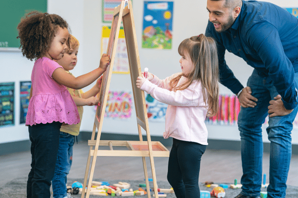 Toddlers drawing on an easel in a preschool classroom, as their teachers looks on