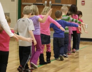 Children int he classroom marching in aline with arms spread wide playing follow the leader game 