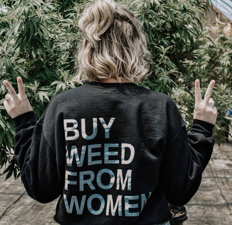Foto colorida de uma mulher de costas, usando um moletom da campanha  “Buy weed from women” - “Compre maconha de mulheres” na frente de um cultivo de maconha