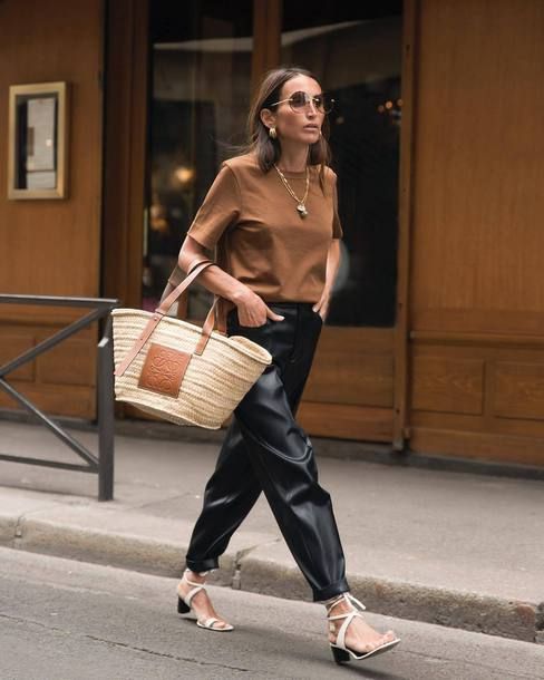 Mulher branca, de cabelos castanhos lisos, usa uma calça cargo e blusa de camurça marrom. Ela está caminhando em uma calçada, utilizando uma bolsa de palha.