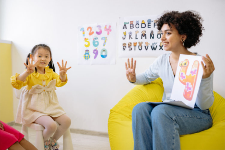 A teacher reading to children from a picture book that is turned to a page with the number four. The teacher and a student hold up four fingers.