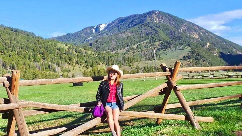 Image of Candy wearing a cowboy hat, sitting on a ranch fence with a mountain in the background