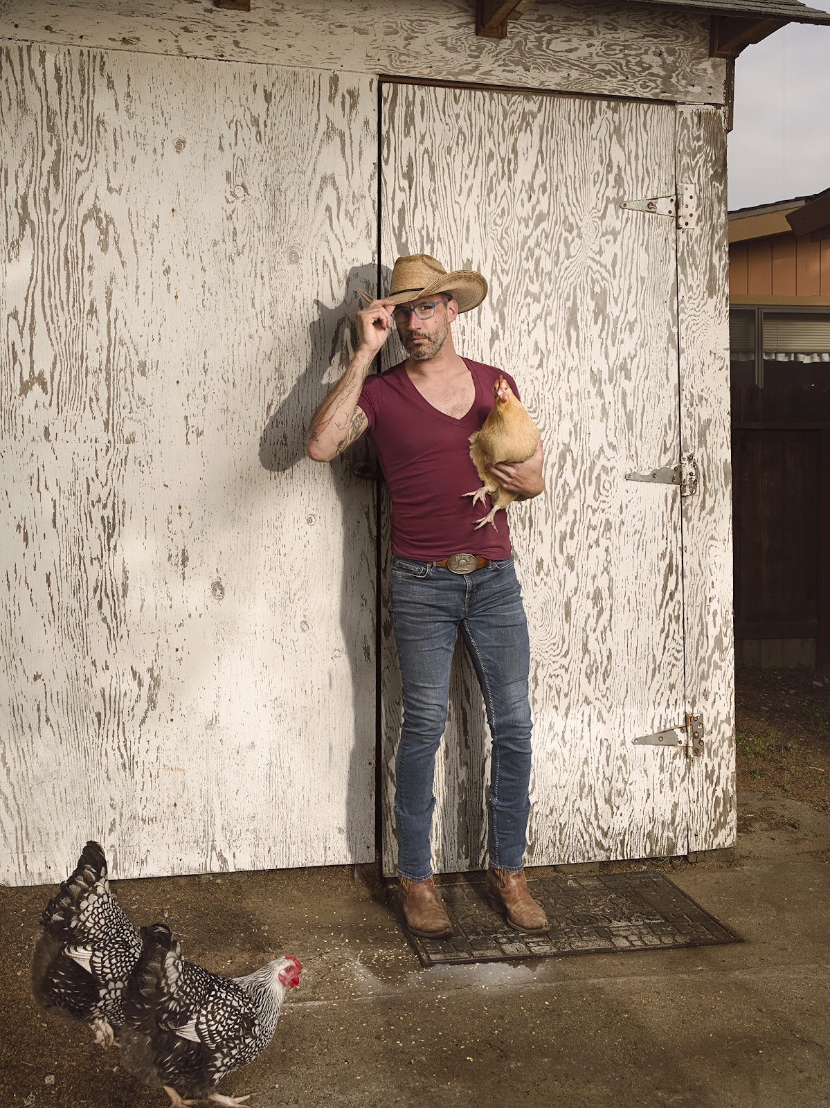 A man stands out against the background of a weathered, white wooden chicken coop. He tilts his stetson down with one hand while holding a caramel-colored hen in the other. A few more chickens are at his feet, pecking at the seeds on the ground.