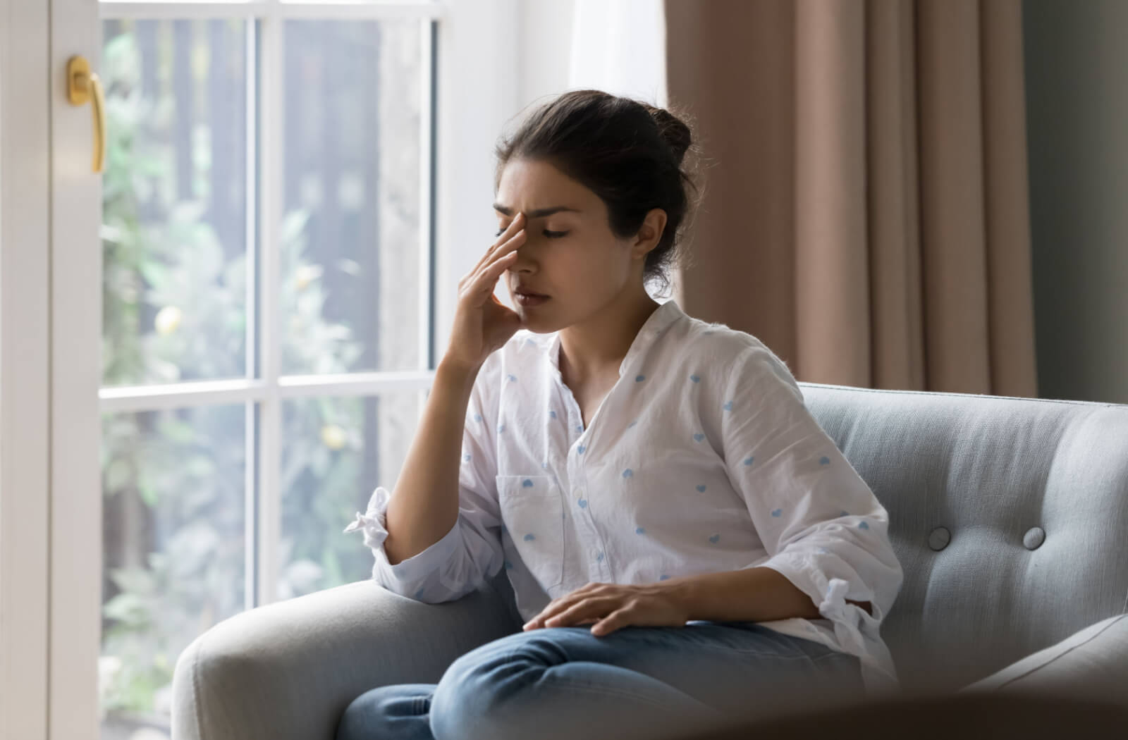 A woman in a brightly lit living room suffering from a headache. Astigmatism-myopia-presbyopia