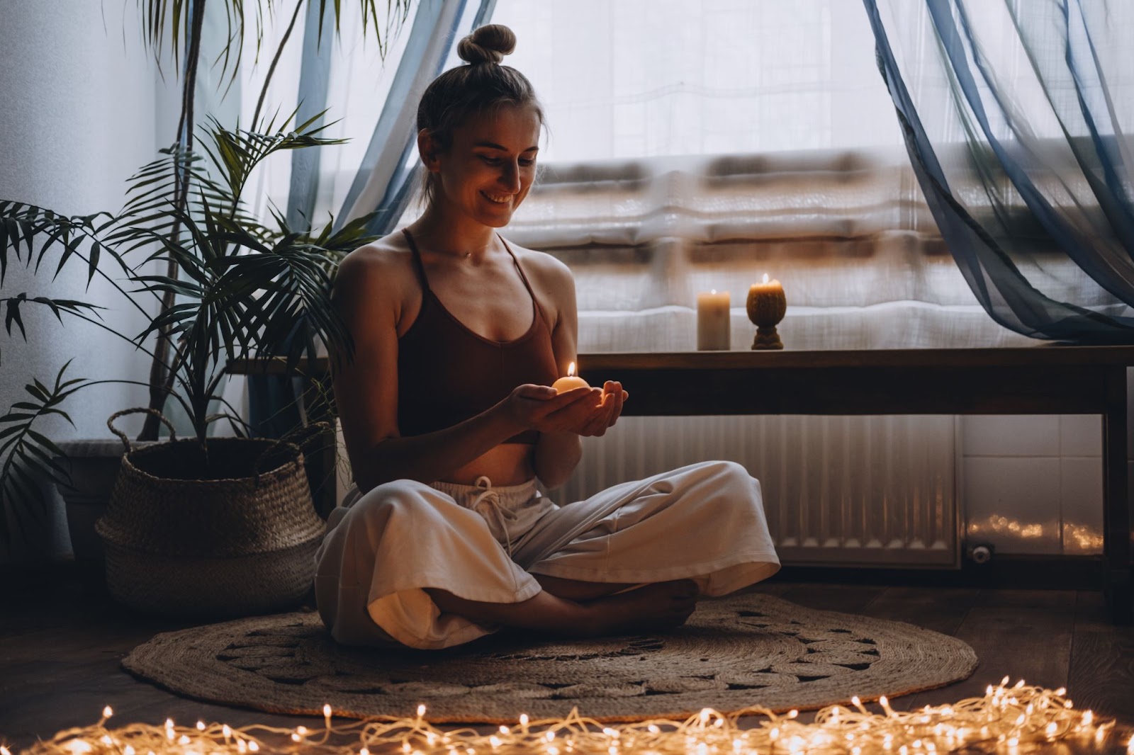 A woman sat on a rug cross-legged, holding a candle in her hand surrounded by fairy lights. 