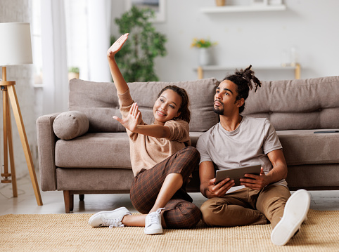 A couple is seen sitting on the floor beside each other. The woman is seated on the left side and is holding her hands out in front of her. The man is seated on the right side next to her holding a tablet in his hands, looking towards the womans hands. Couple is able to communicate. 