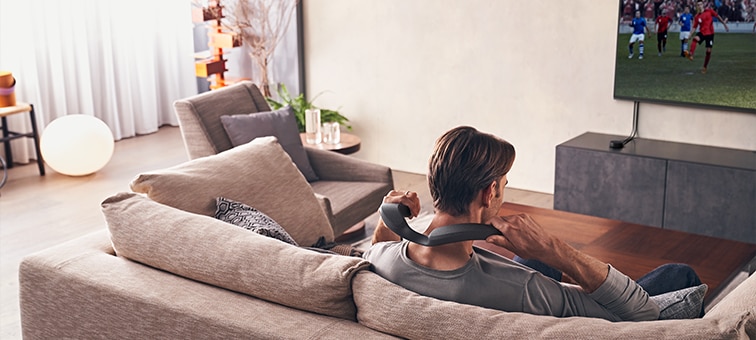 Man sitting on sofa watching film on wall-mounted TV using SRS-NS7 wearable speaker and WLA-NS7 wireless transmitter
