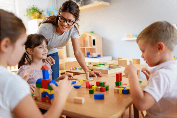 Three children sitting at a table playing with colorful blocks, an adult woman wearing glasses smiling and touching one of them on the shoulder.