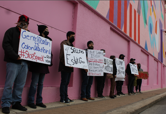 imagen de manifestantes frente a un mural del centro de Brownsville con pancartas que contienen texto sobre detener el aburguesamiento, la colonización y SpaceX (por Gaige Davila)