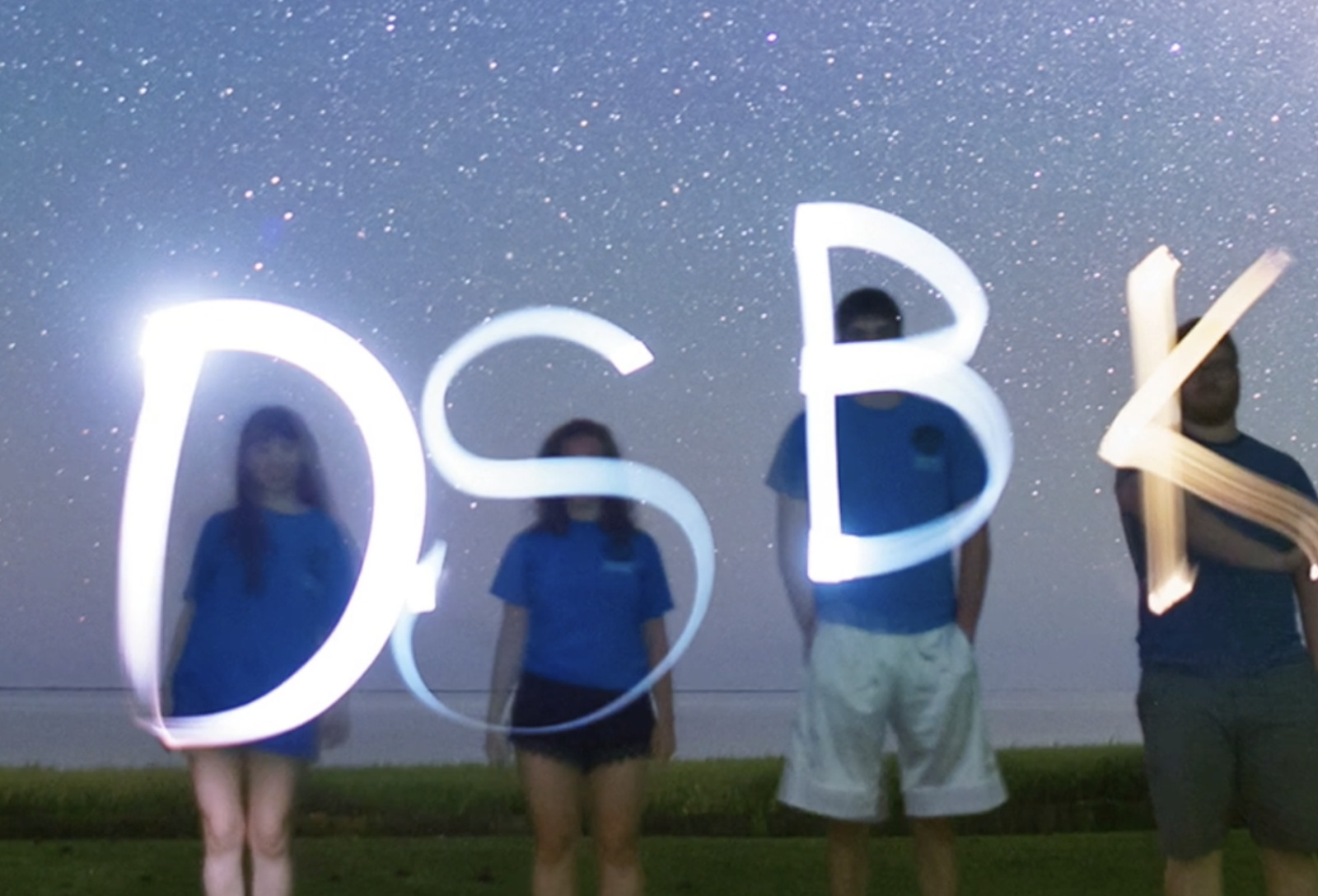 Four people standing outside at night with a starry sky and letters "DSBK" written in light in a long exposure image