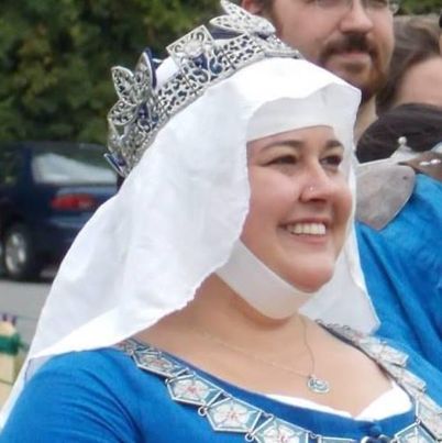 A smiling woman wears a blue dress, enamel collar of roses, a white veil, and a crown.