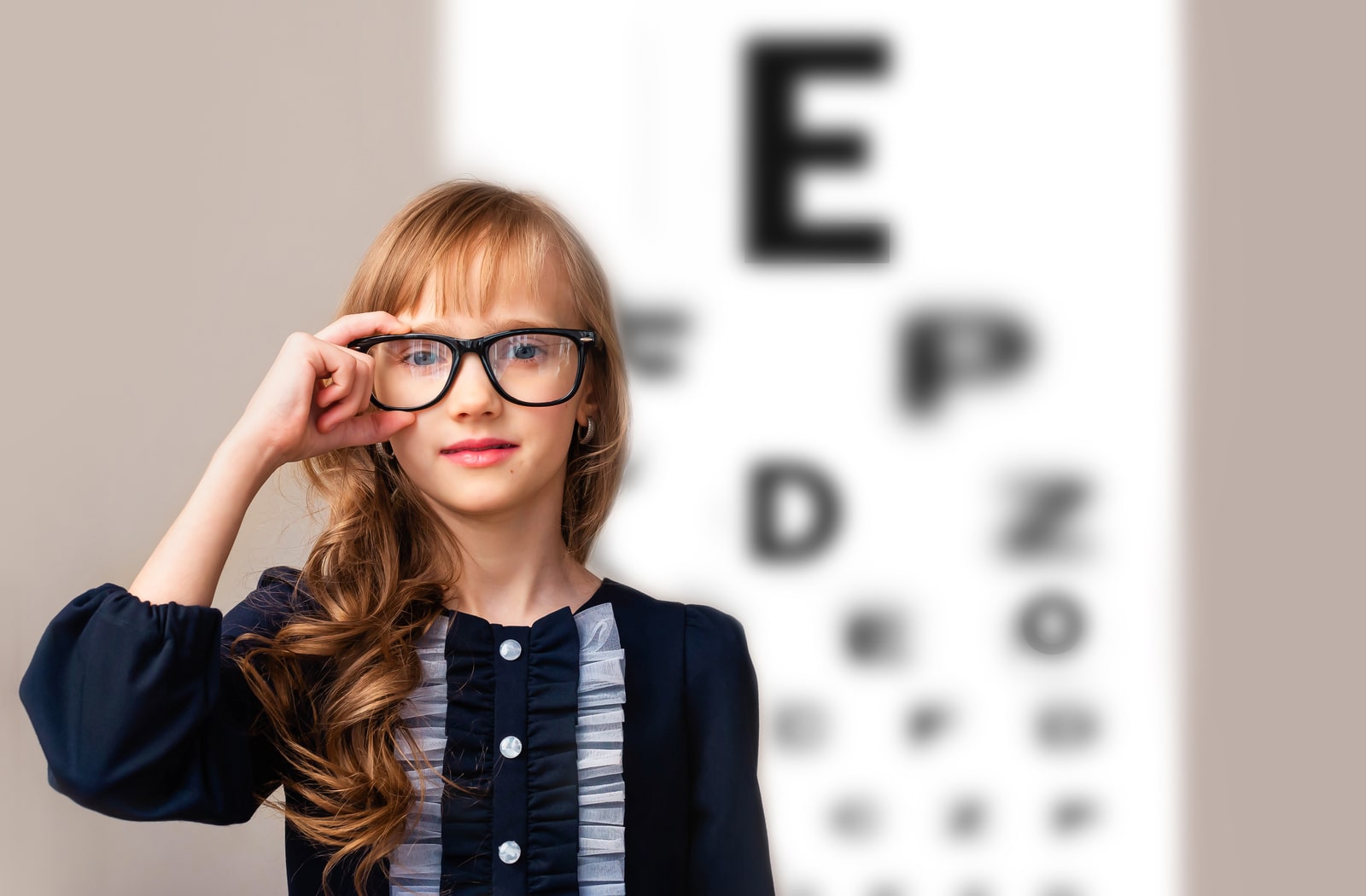 A young girl standing in front of a Snellen eye chart, with her hand on her glasses