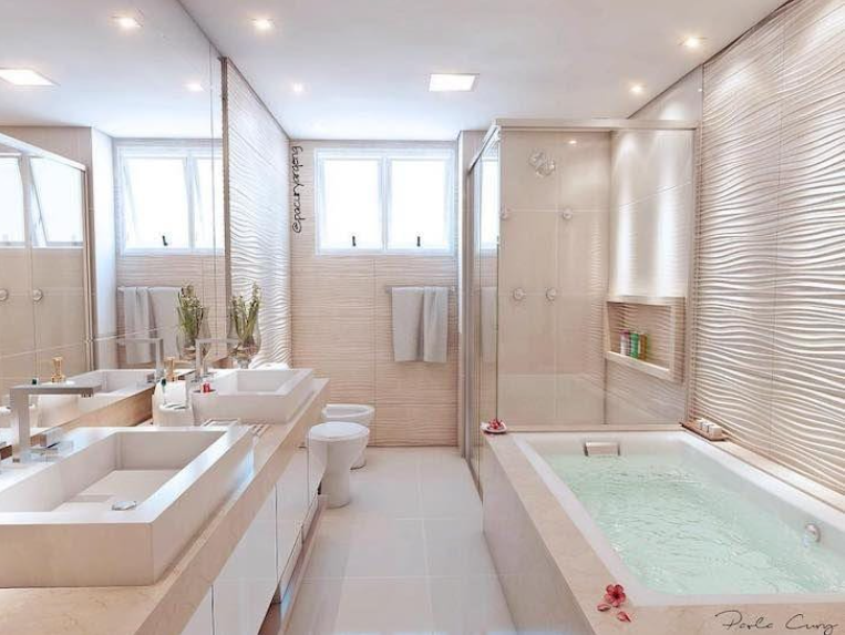 Banheiro com revestimento 3D branco por toda parede, box de vidro, piso branco, banheira embutida, bancada da pia de mármore nude e duas cubas brancas.