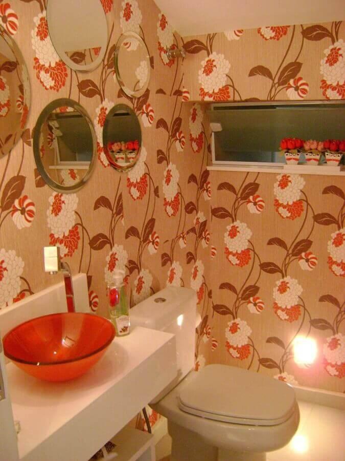 Lavabo em estilo minimalista com papel de parede floral em tons laranjas, cuba de vidro laranja e 4 espelho redondo na parede.