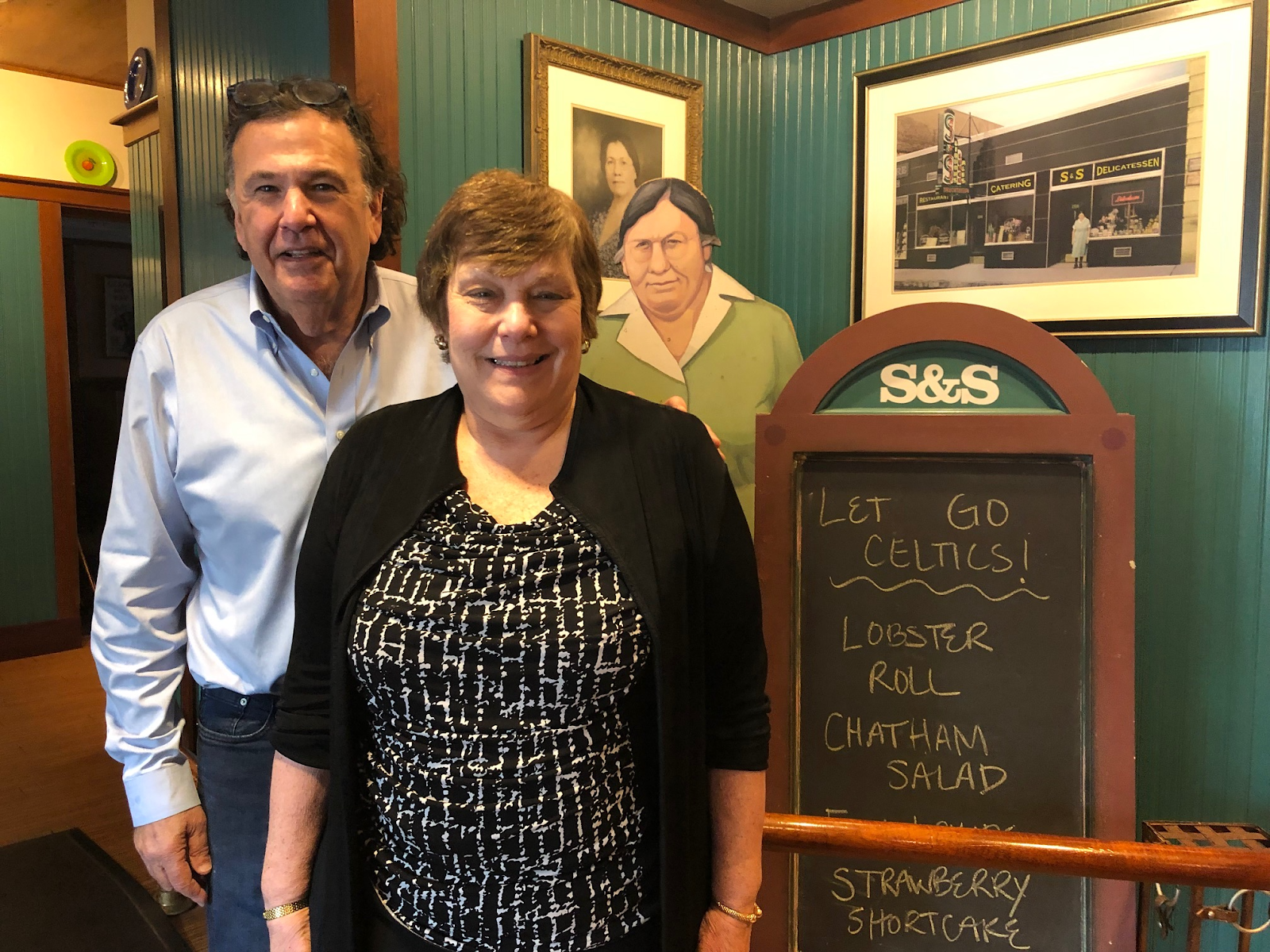 Man with short, dark hair and light blue shirt standing next to a woman with light brown hair and black sweater in front of framed historical photographs. Next to a chalkboard that reads "Let's Go Celtics!/ Lobster Roll/Chatham salad/strawberry shortcake"