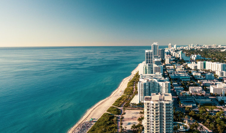 Aerial view of the coast along Miami, Florida. The buildings look a bright white in the sun, the water is a stunning blue, and there’s not a cloud in the sky.