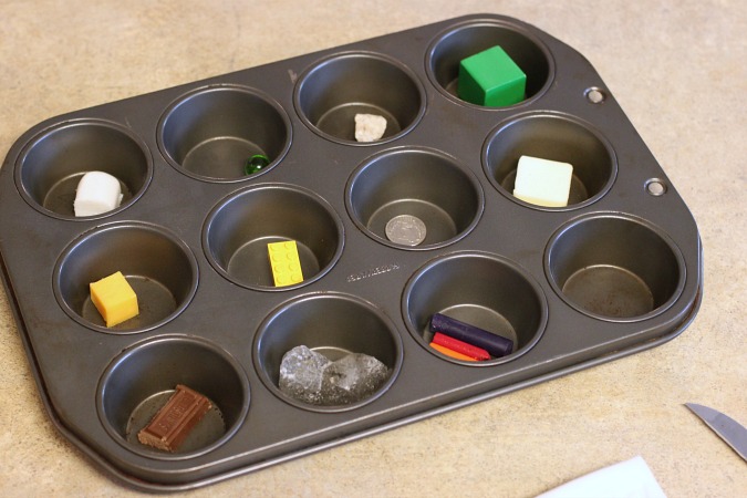 a 12-compartment muffin tin filled with various items including a marshmallow, marble, rock, wooden block, cube of cheese, single lego, coin, square of butter, square of chocolate, ice cube, pieces of crayon.