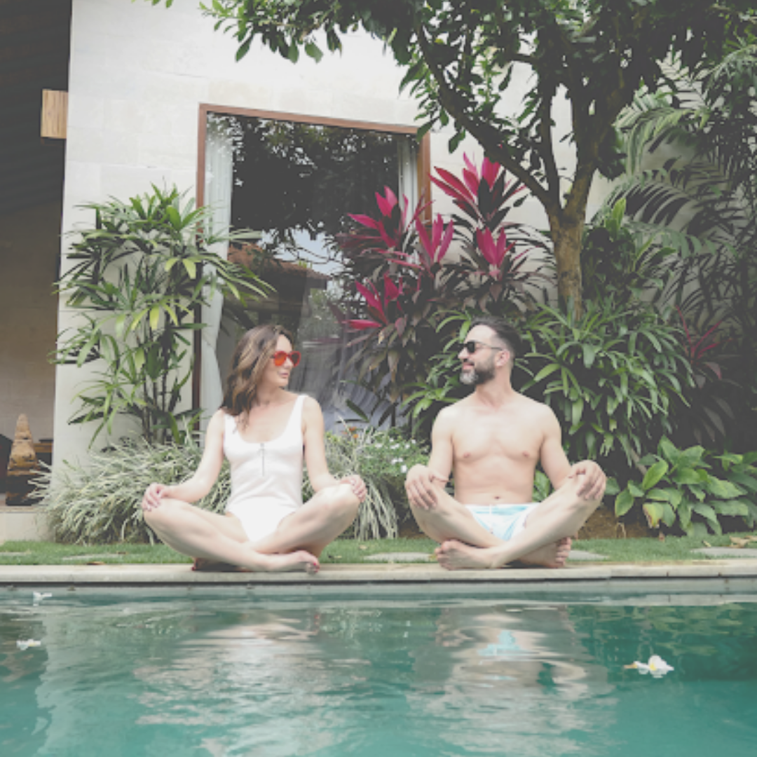 Husband and wife sitting cross-legged beside a pool in bali resort with palm streets and tropical plants around