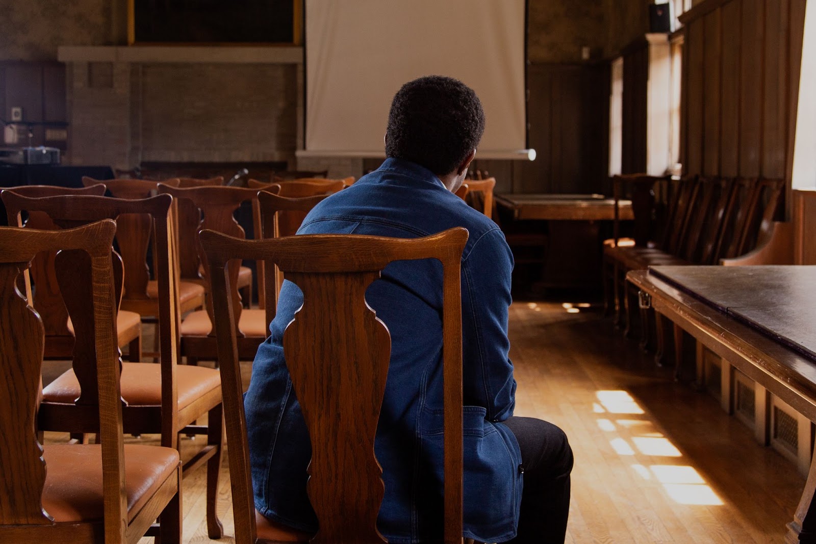 Image: Ross Stanton Jordan sits, back turned to the camera, in a room of the Jane Addams Hull House Museum, again surrounded by variations on the color brown through a wooden table and chairs next to him and wood paneling and wall paper on the walls around him. His head is slightly framed by a projector screen in the distance. He's wearing a denim jacket, dark brown pants. Photo by Joshua Clay Johnson.
