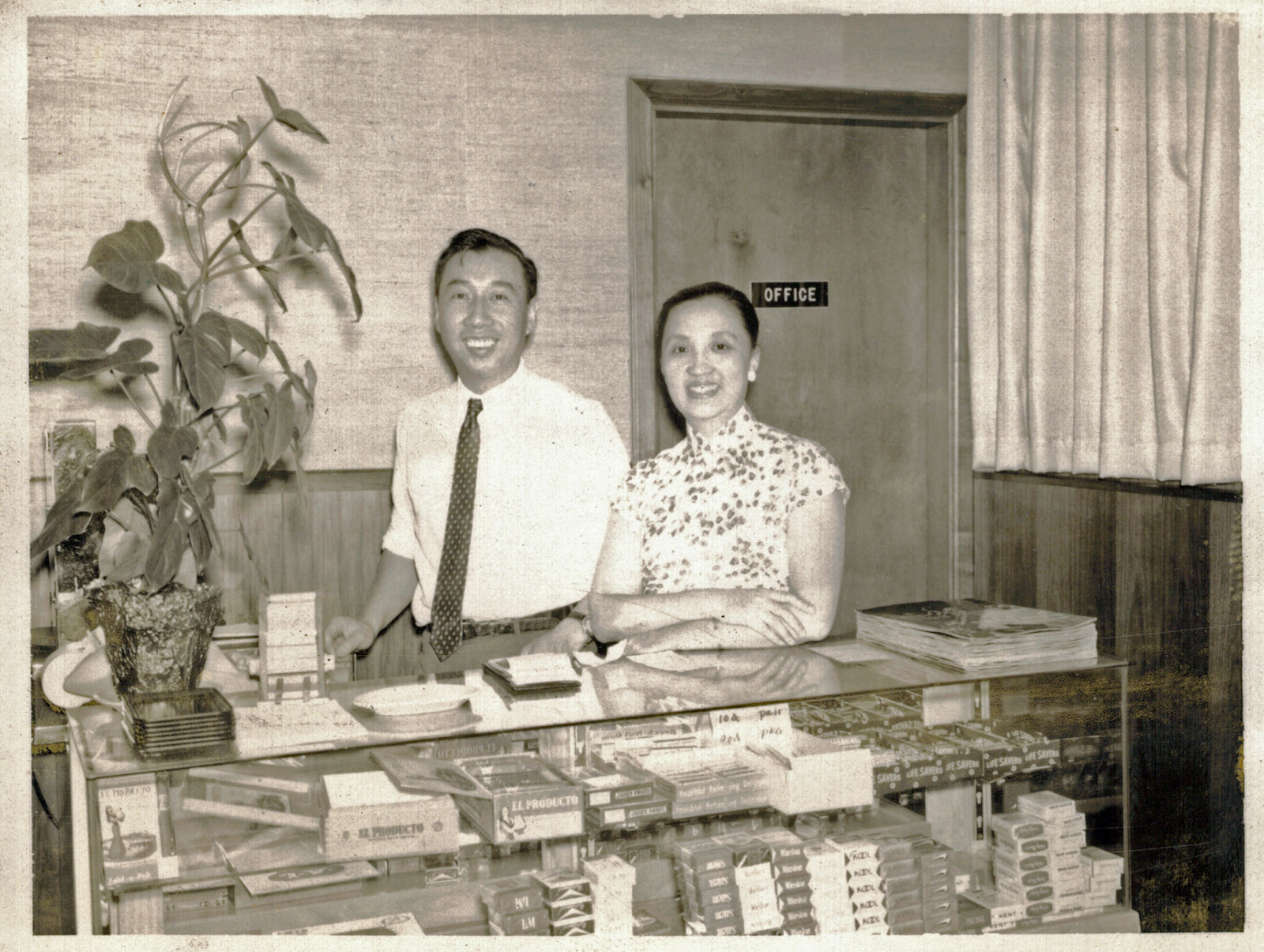 Two smiling people standing behind a glass case. Man on left wears a white white with a dark tie. Woman on right has dark hair and wears a short-sleeved floral dress. In background is a door that reads "office"