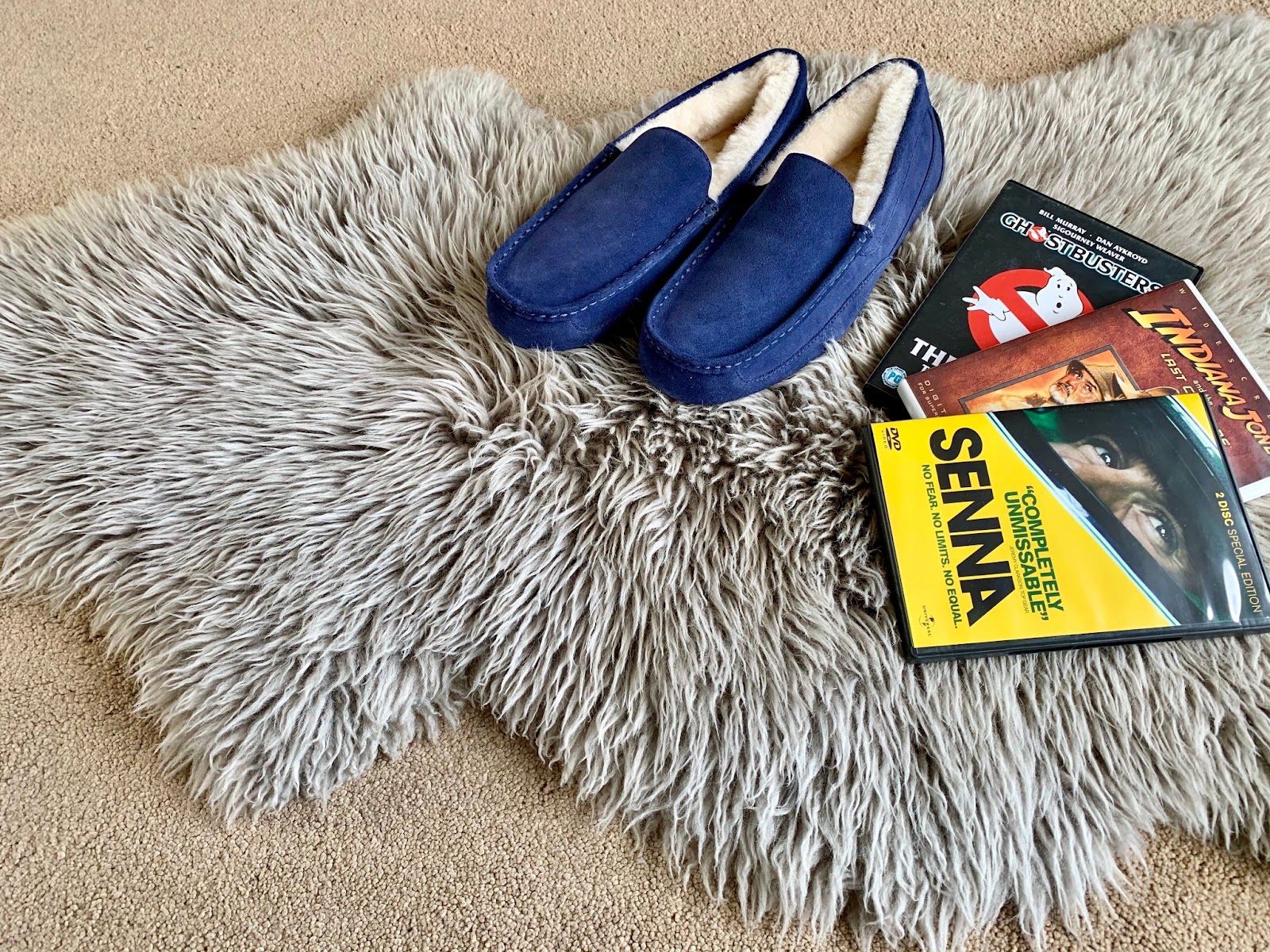 Men's Navy Blue Moccasin slippers sat on a fluffy grey sheepskin rug with 3 DVDs beside them