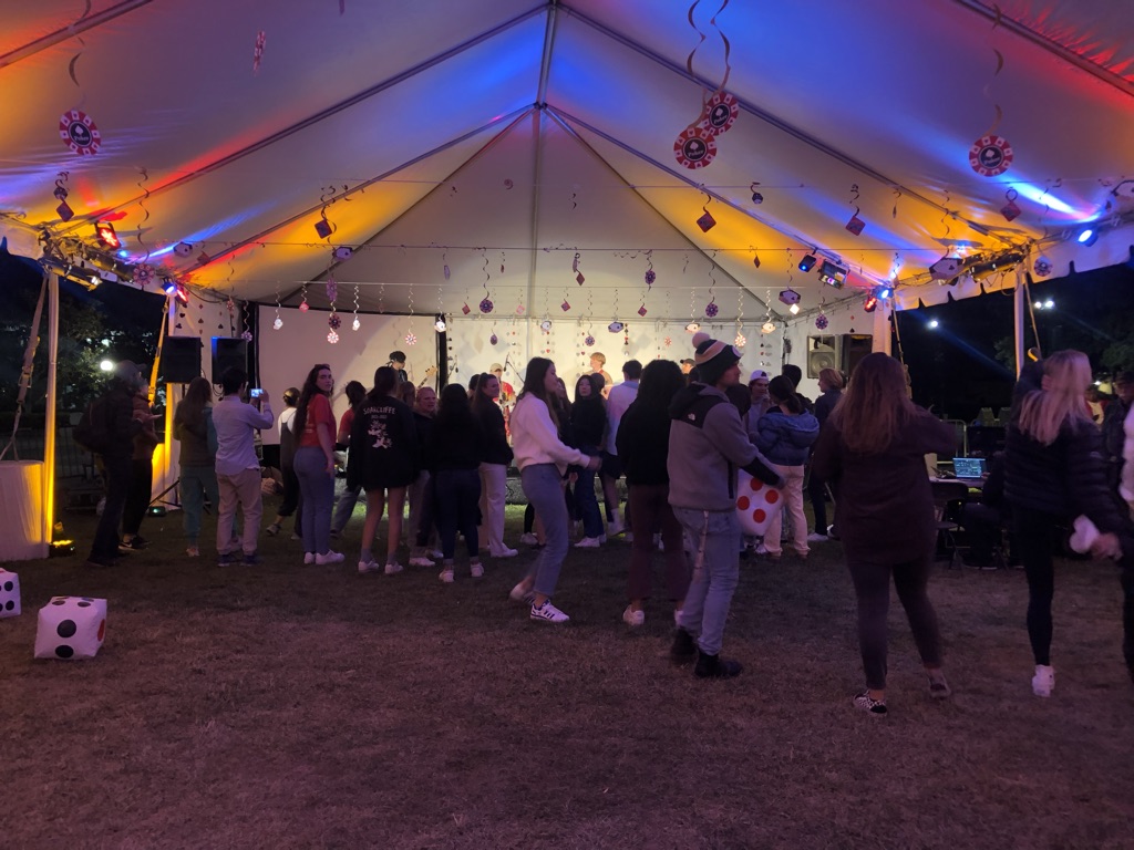 A large tent with orange, blue, and red lighting over a few dozen people dancing and listening to music at night. Stanford band Banana Bred performs under bright white lights behind.
