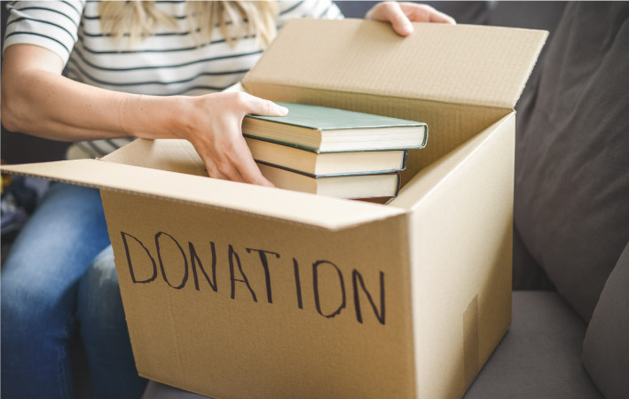 A woman is sitting on her couch, placing books in a box for donation.