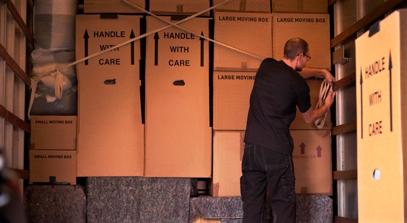 A man uses straps to secure a section of boxes in the back of a moving container. He uses the straps to create an "X" across the section, which will help keep the boxes from moving in transit.