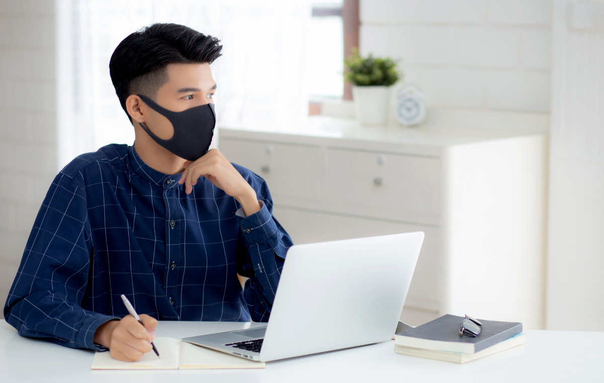 A young student is participating in an online examination at home and is wearing a black face mask.
