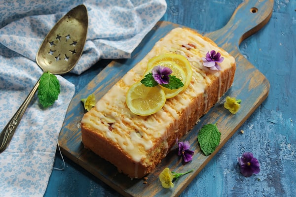 Glazed pound cake with purple flowers and slices of lemon on top, served on a wooden board