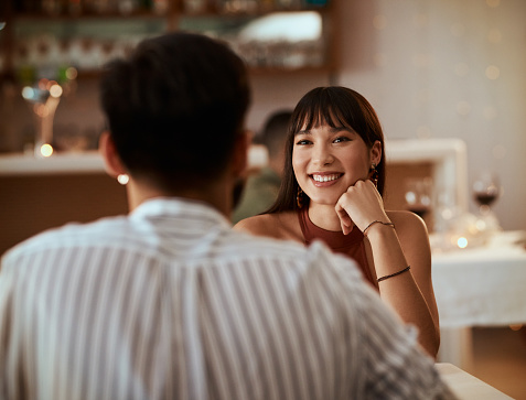 A couple is on a date at a restaurant. They are sitting across from each other, as the woman smiles at him with her hand holding her head up. Communicate with each other. Learn to communicate. 