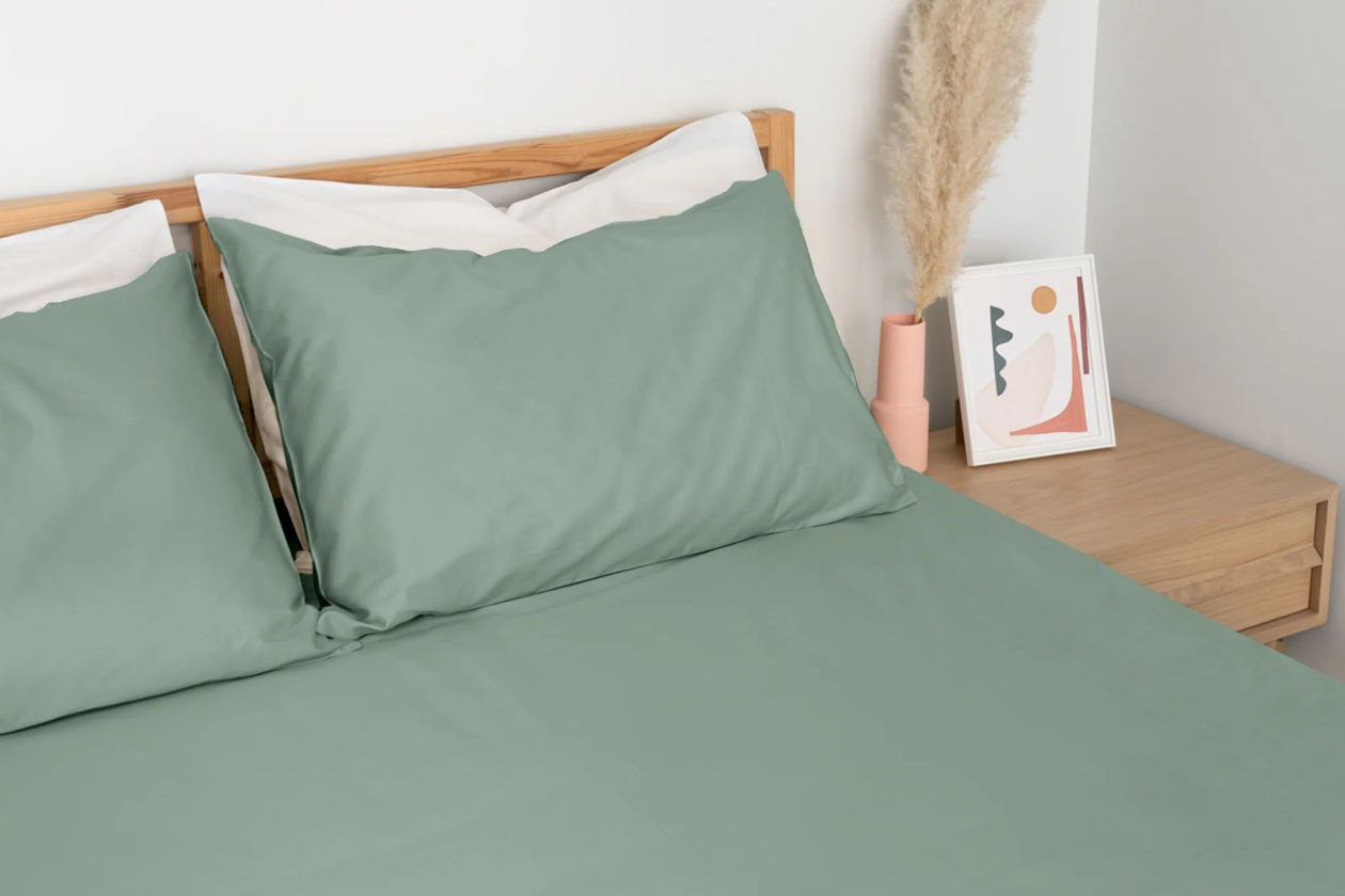 A bed made with Homebird Percale Sheets in sage green. The side table next to the bed holds artwork and a decorative plant.