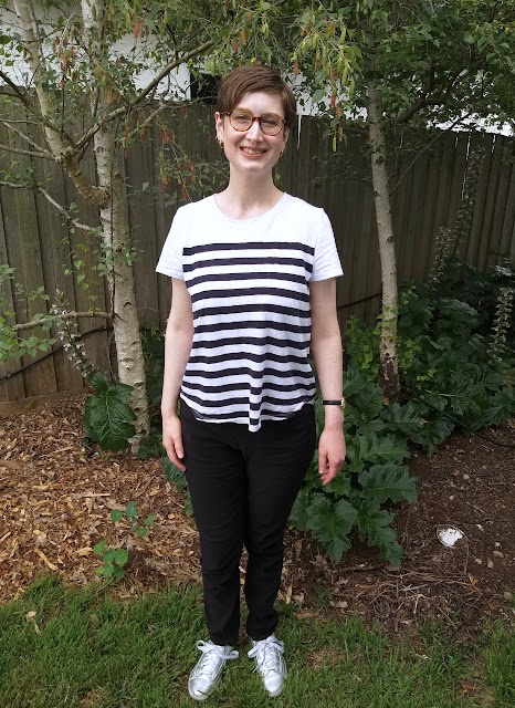 Siobhan stands in front of a garden fence. She wears slim fit black pants with hard-to-notice front pockets, striped white and navy short sleeve tee and silver runners.