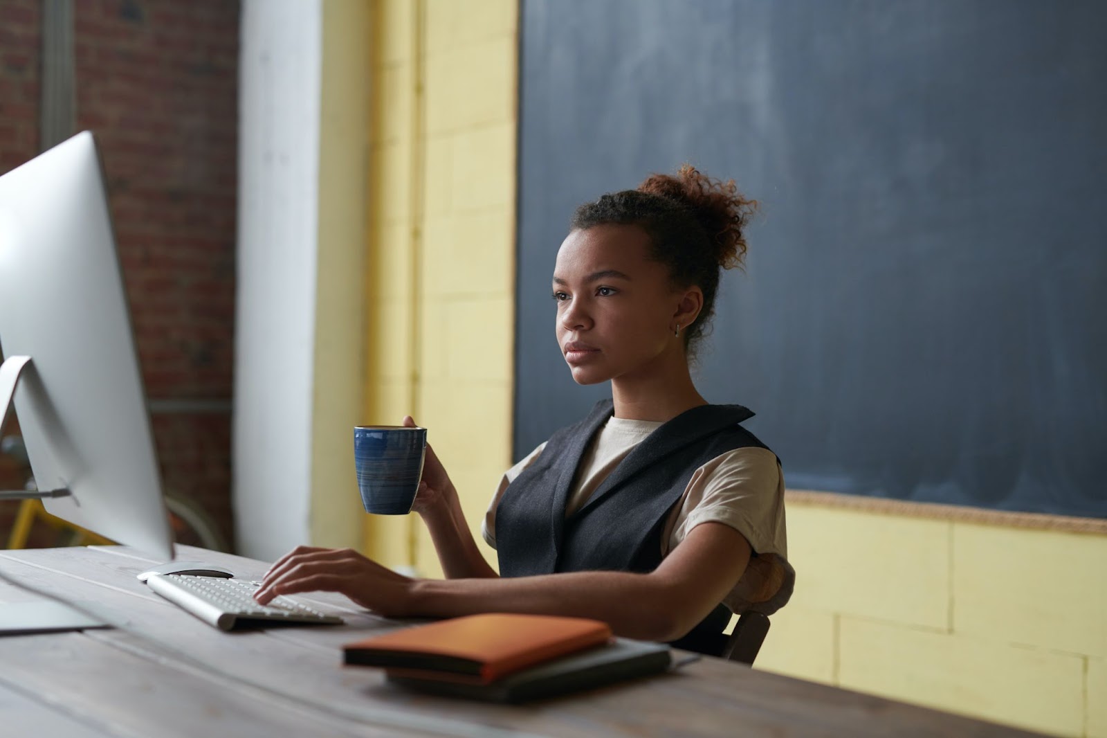 A woman is siting at her desk looking at her computer. She is holding a blue mug in her right hand, and the fingers on  her left hand are on the computer keyboard. An orange book and a blue book are on the desk, and a blackboard on a yellow wall is behind the woman.