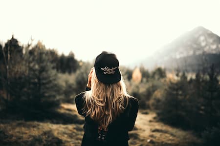 Une femme blonde portant une casquette noire est de dos et regarde un décor montagneux boisé.