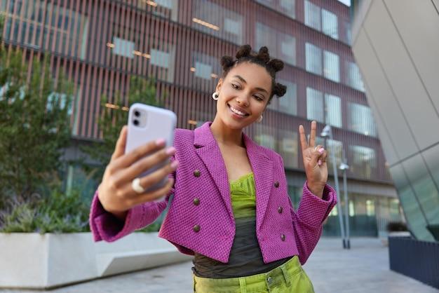 Attractive girl in fashionable outfit creats influence content shows peace sign at smartphone front camera poses for selfie smiles gladfully poses at urban place spends vacation at big city. Free Photo