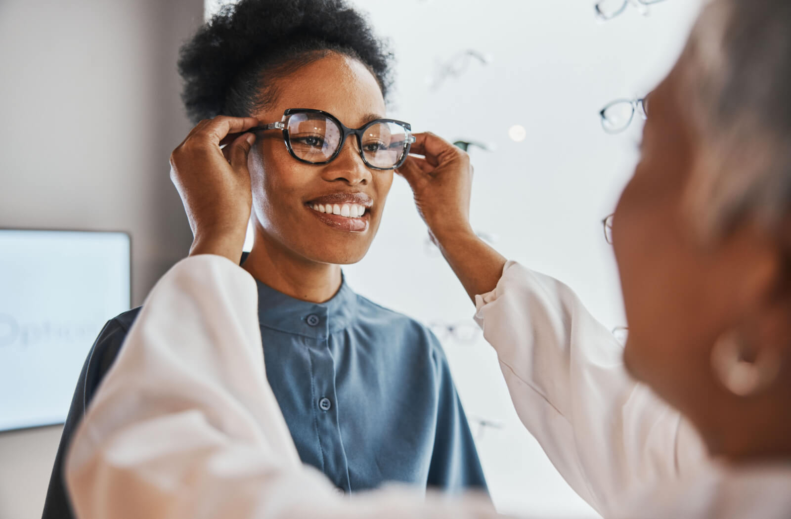 A young woman smiling and trying on glasses in a store while being assisted by an optometrist