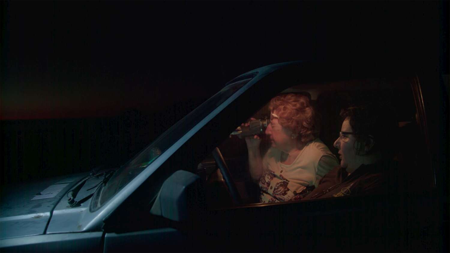 Silvia and Andrea sit in their car at night. The overhead light is on, as they peer outside with binoculars.