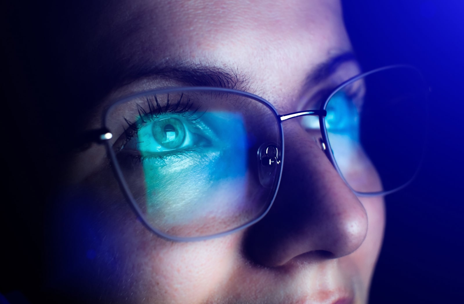 Close-up of a woman wearing glasses in a dark room with blue light from a screen reflecting off her glasses
