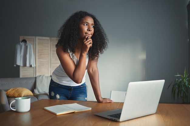 Picture of thoughtful dark skinned student girl in tank top and jeans touching chin with pensive facial expression, holding pen while writing essay, doing homework, standing in front of open laptop